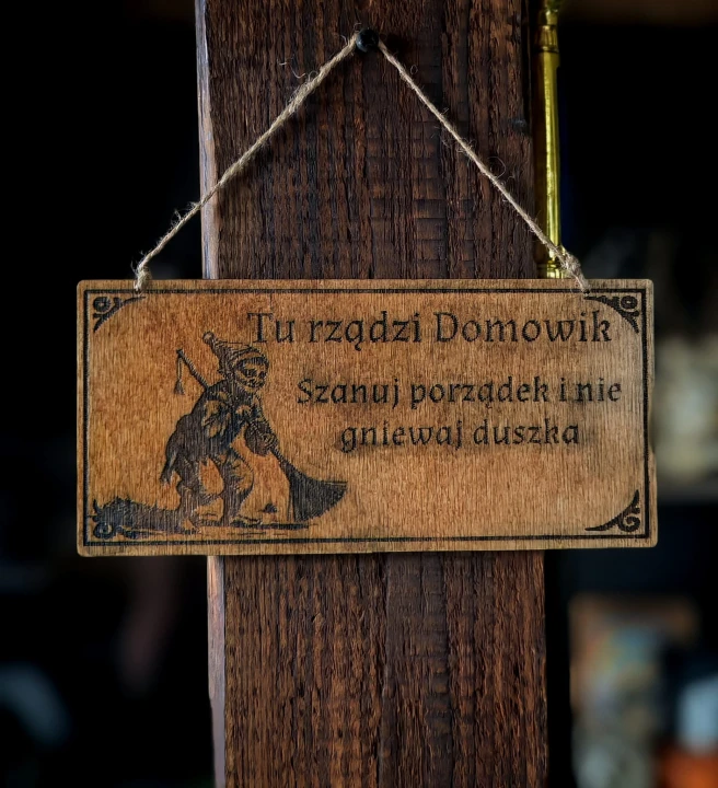 Dekoracyjna grawerowana tabliczka ze słowiańskim napisem  „Tu rządzi Domowik. Szanuj porządek i nie gniewaj duszka” –  ozdoba do domu inspirowana folklorem i słowiańskim humorem.