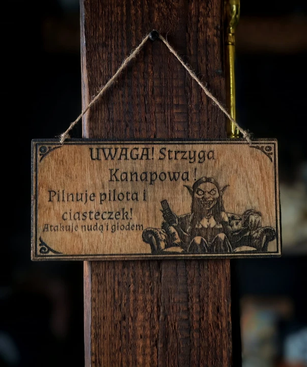 Dekoracyjna grawerowana tabliczka ze słowiańskim napisem „Uwaga! Strzyga Kanapowa! Pilnuje pilota i ciasteczek!” –  ozdoba do domu inspirowana folklorem i słowiańskim humorem.
