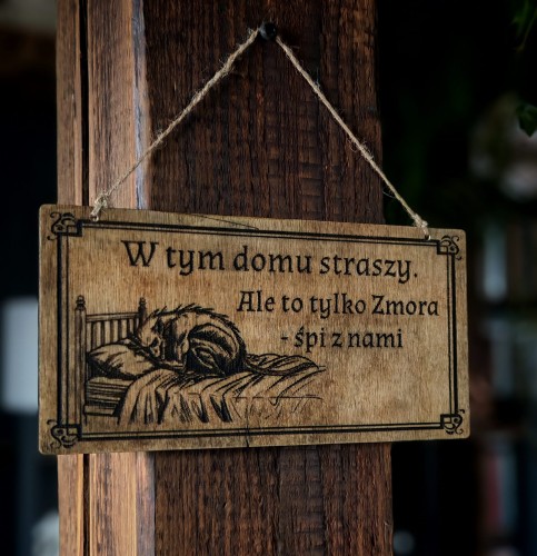 Dekoracyjna grawerowana tabliczka ze słowiańskim napisem „W tym domu straszy.  Ale to tylko Zmora- śpi z nami.” –  ozdoba do domu inspirowana folklorem i słowiańskim humorem.