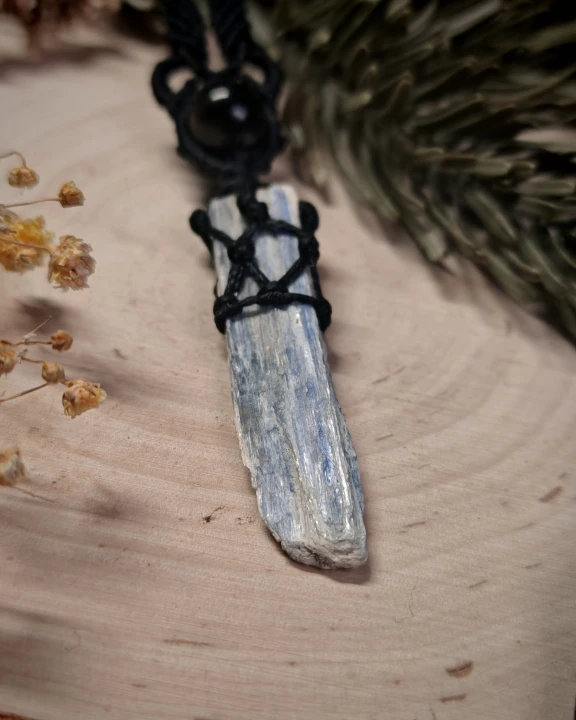Ręcznie wykonany wisiorek z naturalnym kyanitem, opleciony czarnym sznurkiem w stylu makramy. Surowy, podłużny kamień w jasnych, srebrzysto-niebieskich tonach pięknie kontrastuje z głęboką czernią plecionki oraz okrągłym, czarnym koralikiem.
